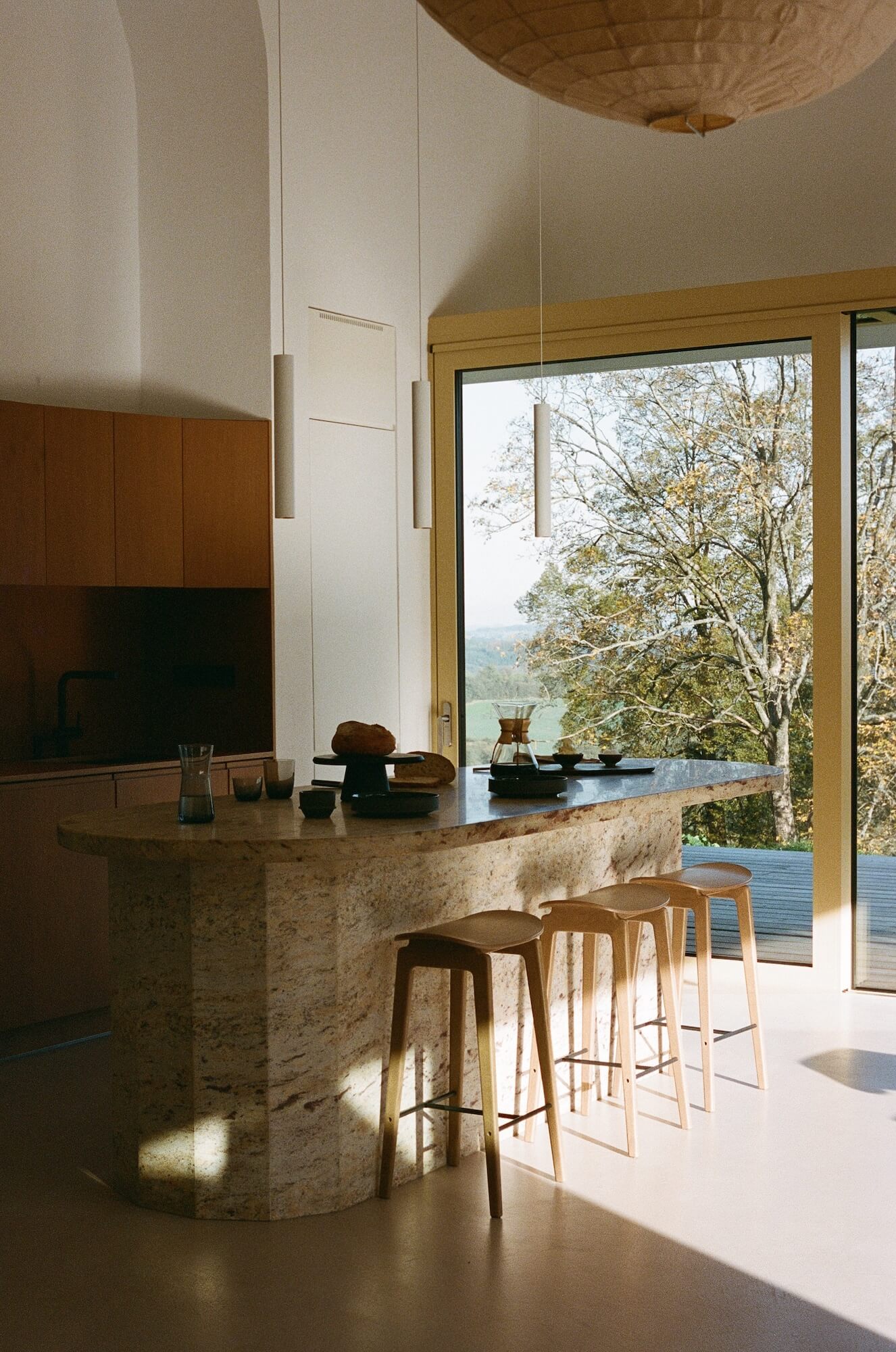 Modern kitchen with a stone island and stools, large windows showing trees outside.